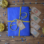 Flat lay of cross stitched gift tags next to a gift with the Santa tag tied to the gift with yellow ribbon