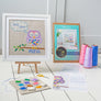 Finished cross stitch pattern shown in a frame with the kit materials including packaging, threads, aida, pattern and needle
