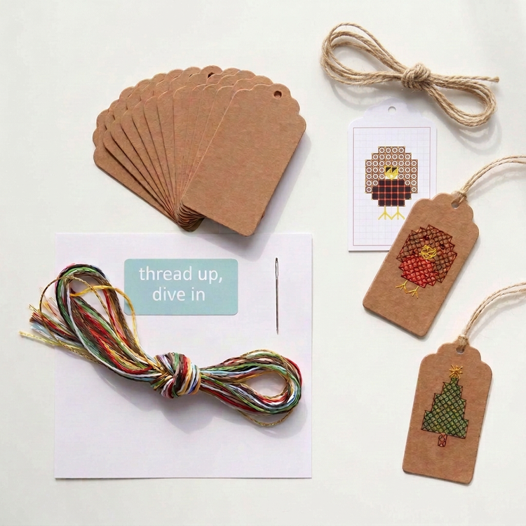 Contents of the Christmas cross stitch gift tag kit: 10 kraft tags, robin pattern, knotted DMC threads, needle and jute twine, with finished robin and tree tags