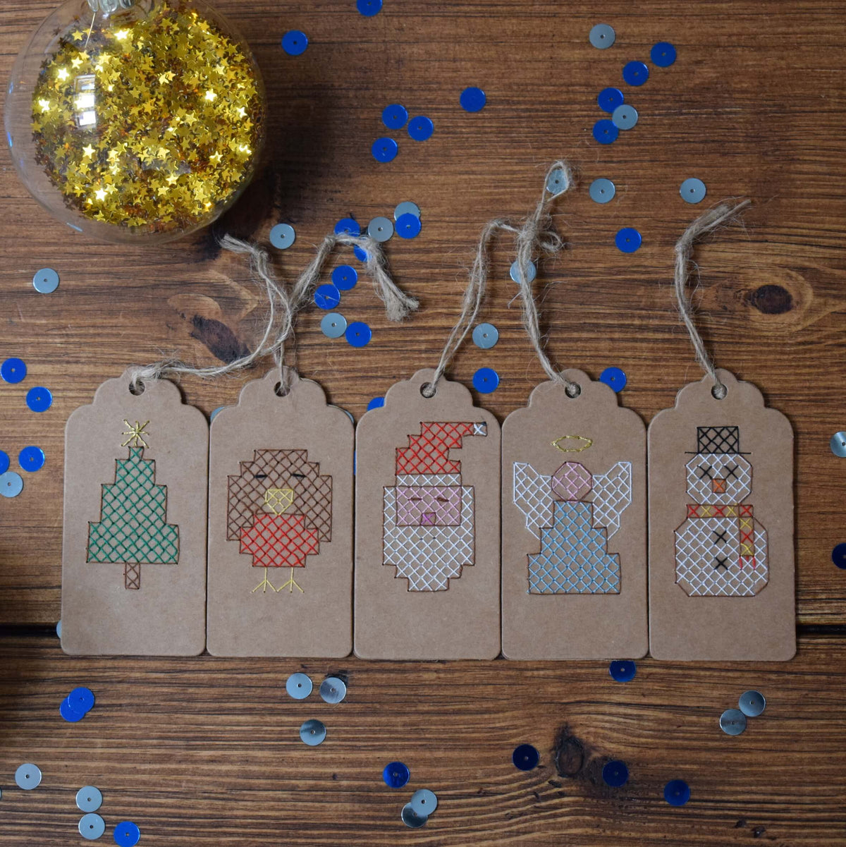 All five cross stitched tags featuring a Christmas tree, robin, angel, Santa and snowman laid side by side on a table