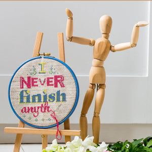 Cross stitch kit pattern finished and framed in a hoop featuring the I never finish anything slogan with a shrugging mini mannequin