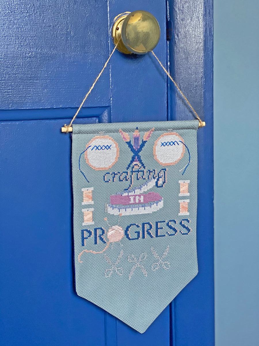 Cross stitch banner featuring crafting on progress slogan hanging on a blue door