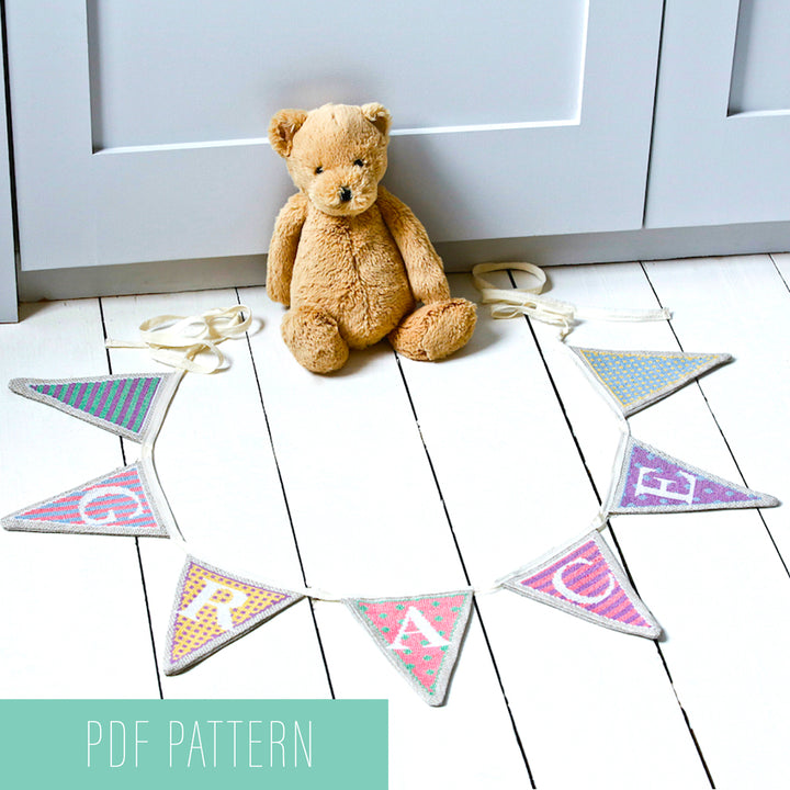 Pastel bunting cross stitch flags spelling grace on floor around teddy