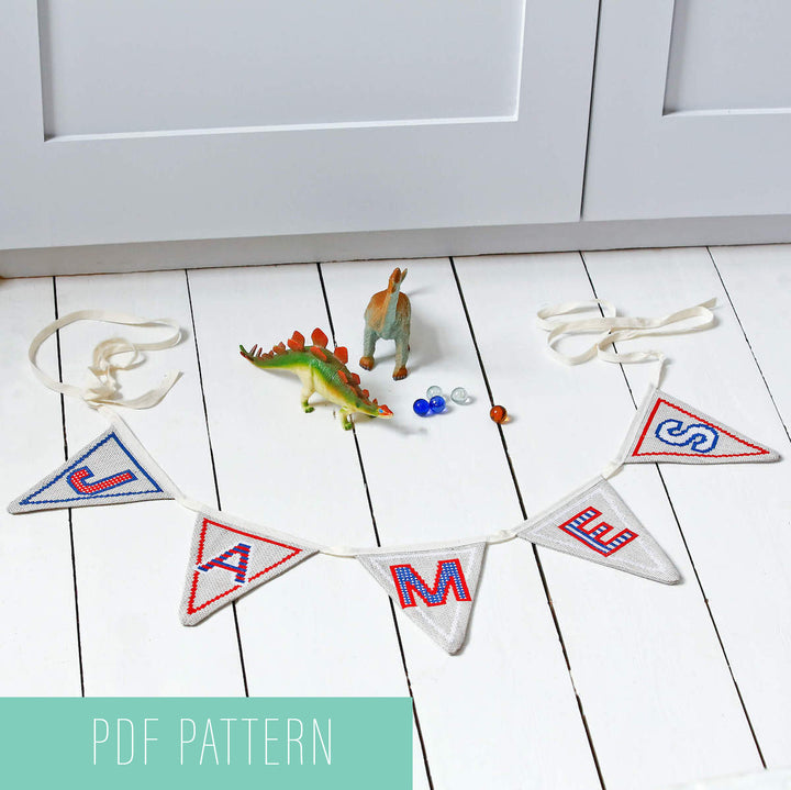 Cross stitch alphabet bunting spells out James on the floor around toy dinosaurs