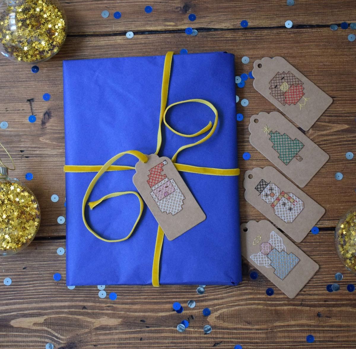 Flat lay of cross stitched gift tags next to a gift with the Santa tag tied to the gift with yellow ribbon