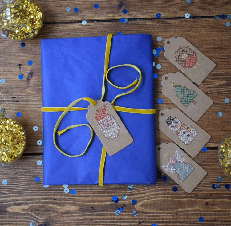 Flat lay of cross stitched gift tags next to a gift with the Santa tag tied to the gift with yellow ribbon