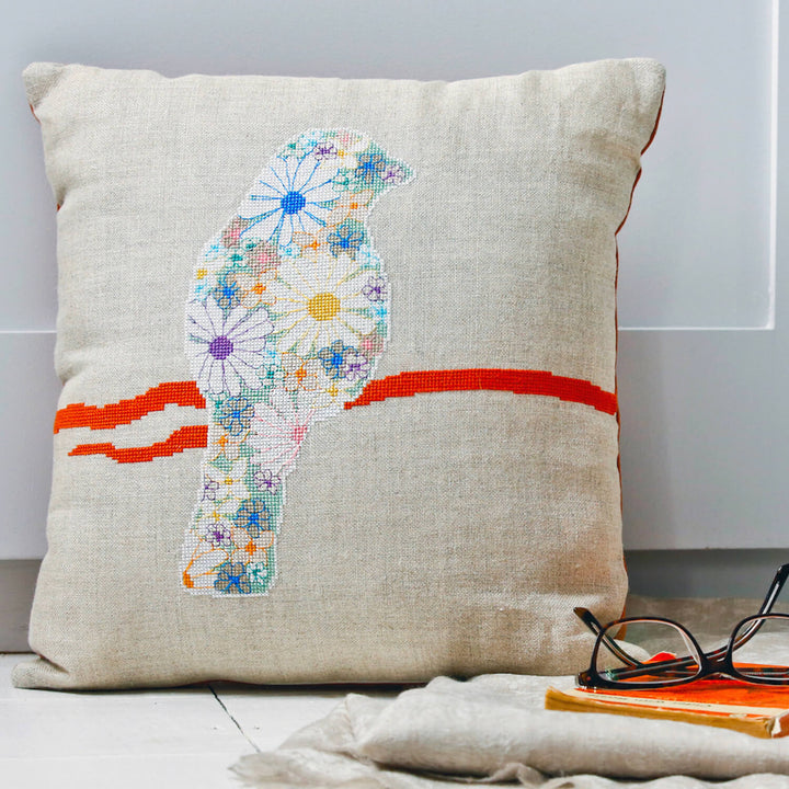 Flower cross stitch bird kit shown made into a cushion shown with a book, scarf and glasses