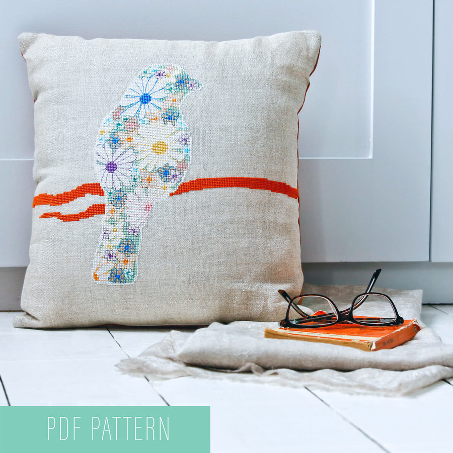 Flower filled bird cross stitch design made into a cushion shown with books and scarf