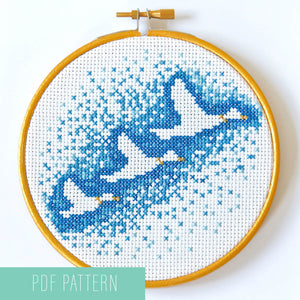 Embroidery hoop containing the outline of three flying ducks surrounded by gradually fading dark to light blue cross stitches.
