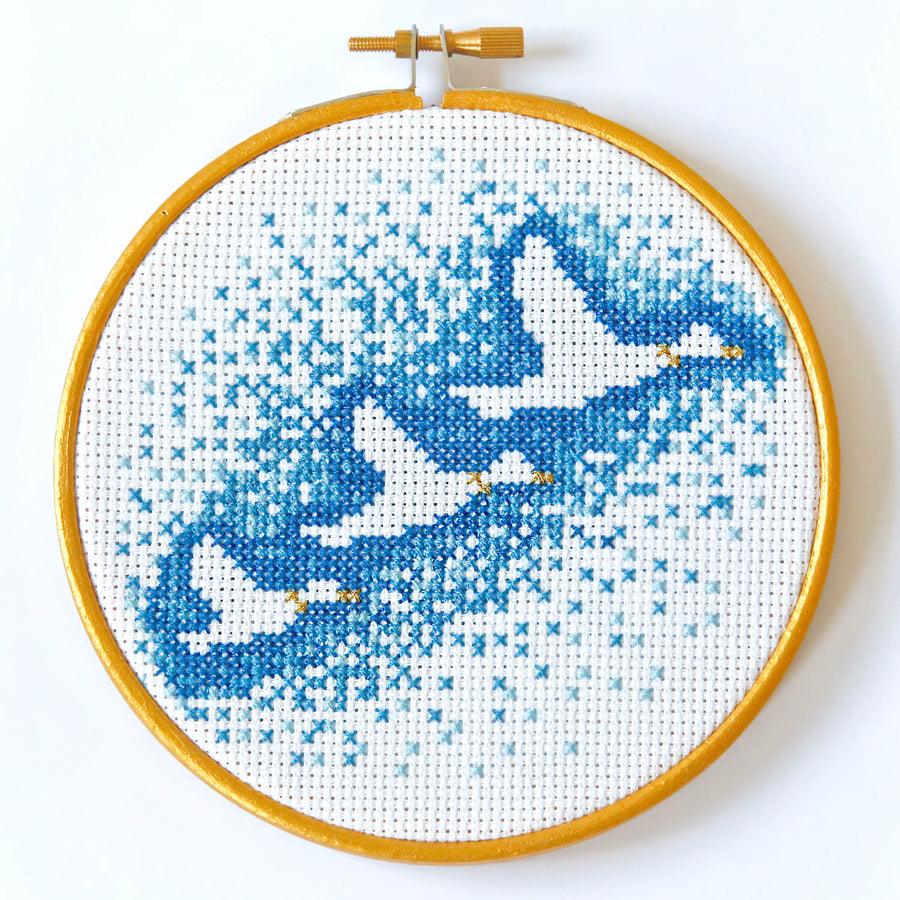 Embroidery hoop containing the outline of three flying ducks surrounded by gradually fading dark to light blue cross stitches.