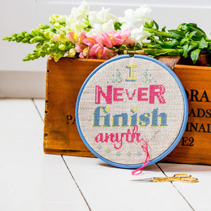 Embroidery hoop filled with slogan saying I never finish anything in pinks and blue/grey stitches, leaning on a box with flowers