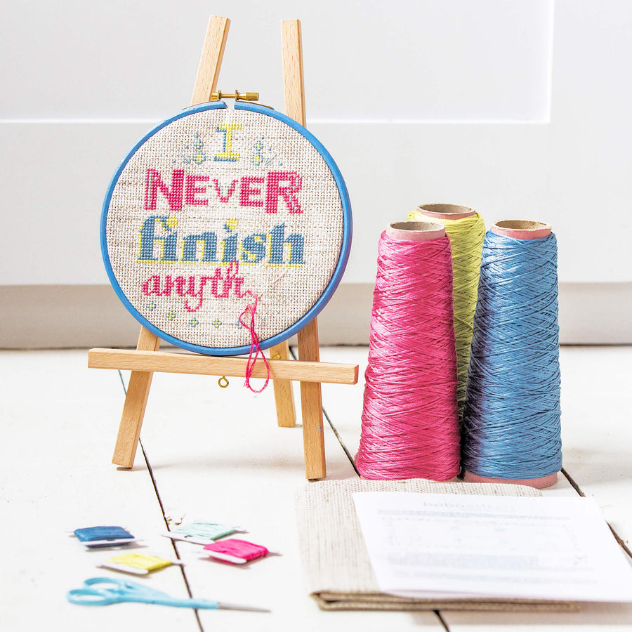 I never finish anythi... stitched embroidery hoop displayed on a mini easel with DMC thread cones, fabric and pattern.