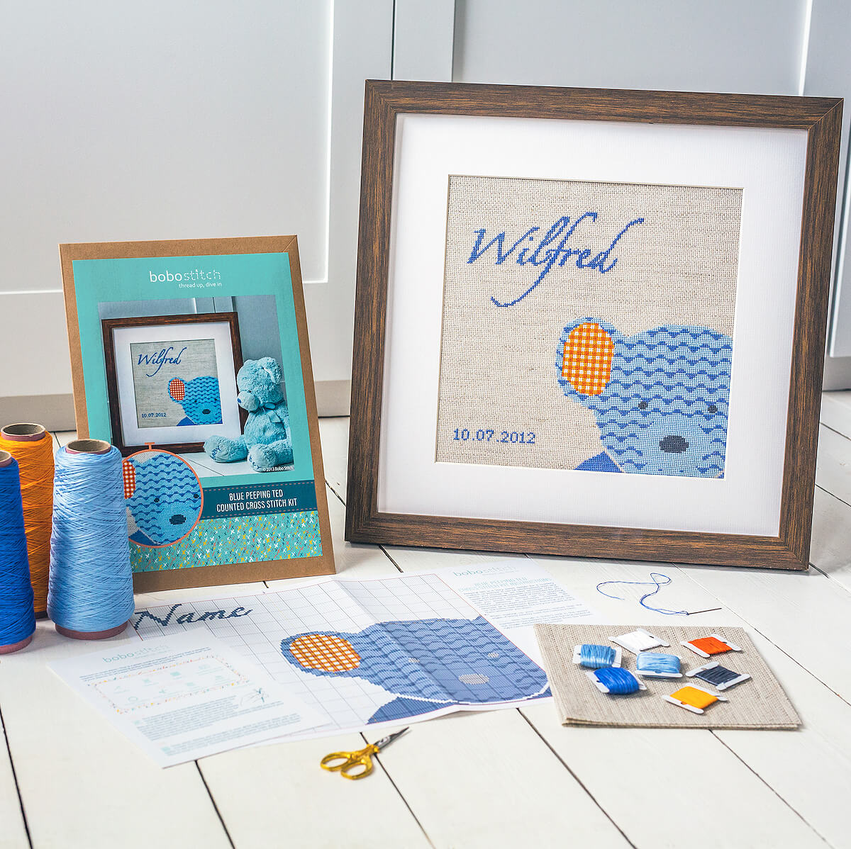 Blue teddy cross stitch design shown framed with threads, pattern and fabric
