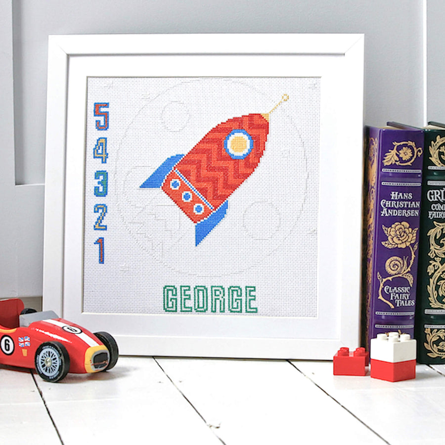 Glow in the dark rocket cross stitch pattern show in a white frame with toys and books