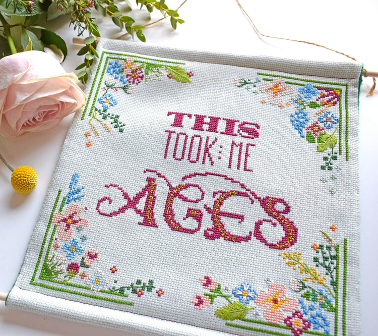 Banner which says This Took me Ages in cross stitch surrounded by flowers