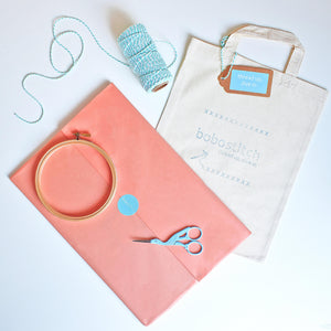 Cross stitch kit wrapped in peach tissue paper with blue stork scissors, small embroidery hoop and cotton bag with gift tag